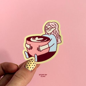 Könnte beinhalten: Ein gestanzter Aufkleber mit einer Cartoon-Illustration einer Person mit langem Haar und Brille, die ein hellblaues Oberteil und eine dunkle Hose trägt und eine große rosa Tasse mit Kaffee und einem weißen Herz-Latte-Art-Motiv umarmt.