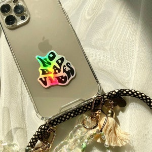 以下が含まれることがあります： ホログラフィック「NO BAD VIBES」ステッカーが付いた透明な携帯電話ケース。 電話は、クリーム色のタッセルとビーズのアクセントが付いた黒と金のロープストラップに取り付けられています。 ケースは白いテクスチャのある表面にあります。