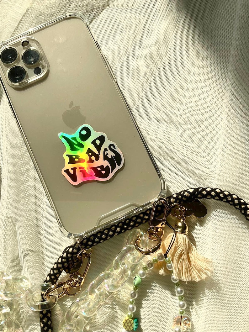 以下が含まれることがあります： ホログラフィック「NO BAD VIBES」ステッカーが付いた透明な携帯電話ケース。 電話は、クリーム色のタッセルとビーズのアクセントが付いた黒と金のロープストラップに取り付けられています。 ケースは白いテクスチャのある表面にあります。