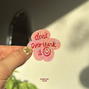 Könnte beinhalten: Ein rosa, blütenförmiger Aufkleber mit dem Text "don't overthink it" in roter Schrift und einem Smiley. Der Aufkleber hat Glitzerakzente und wird von einer Hand mit einem goldenen Stern-Nageldesign gehalten. Die Worte "STUFF BY CLAU" stehen unten.