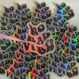 以下が含まれることがあります： 黒い波状のフォントで「NO BAD VIBES」というフレーズが書かれたホログラフィックステッカーのコレクション。ステッカーは反射性のある虹色の仕上がりです。ピンセットが1つのステッカーを持っています。白い表面に散らばっています。