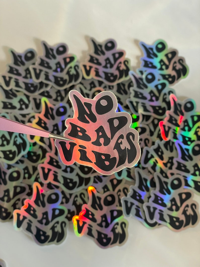 以下が含まれることがあります： 黒い波状のフォントで「NO BAD VIBES」というフレーズが書かれたホログラフィックステッカーのコレクション。ステッカーは反射性のある虹色の仕上がりです。ピンセットが1つのステッカーを持っています。白い表面に散らばっています。