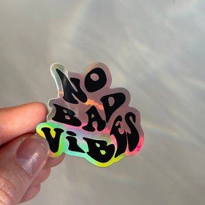 以下が含まれることがあります： 黒いスタイルの文字で「NO BAD VIBES」と書かれたホログラフィックステッカー。ステッカーは虹色のイリデセント効果があり、さまざまな色を反射します。ステッカーは人の手で持たれています。