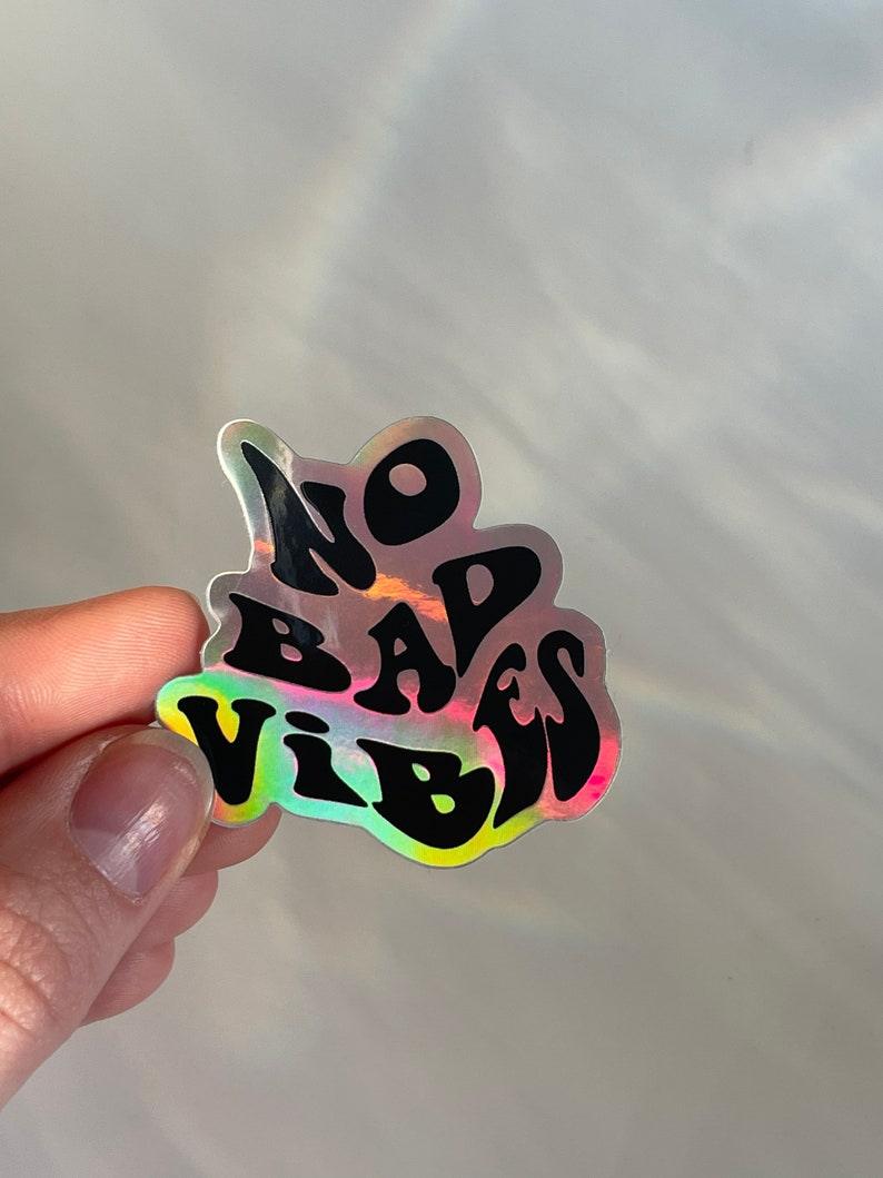 以下が含まれることがあります： 黒いスタイルの文字で「NO BAD VIBES」と書かれたホログラフィックステッカー。ステッカーは虹色のイリデセント効果があり、さまざまな色を反射します。ステッカーは人の手で持たれています。