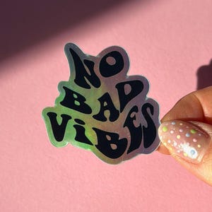 以下が含まれることがあります： 黒いスタイルの文字で「NO BAD VIBES」と書かれたホログラフィックステッカー。ステッカーは虹色の光沢があり、ピンク色の背景に配置されています。