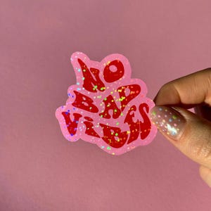 Könnte beinhalten: Ein pinkfarbener Aufkleber mit den Worten "NO BAD VIBES" in roten, glitzernden Buchstaben. Der Aufkleber hat einen holografischen Effekt und wird von einer Hand gehalten.