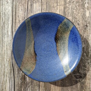 May include: A round ceramic bowl with a blue glaze and tan and brown streaks. The bowl is handmade. The bowl is on a wooden surface. The diameter of the bowl is approximately 15 cm.