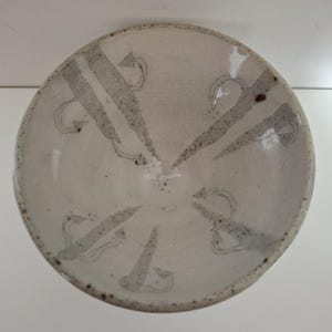May include: A round ceramic bowl with a speckled off-white glaze and a fine crackle finish. The interior features a radial pattern of dark grey, brush-stroked triangular shapes and thin, curved lines that extend from the centre to the rim.