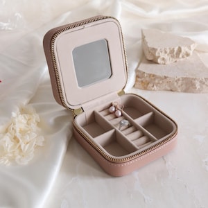 May include: A small, open jewellery box in blush pink, with a gold zip and a mirror inside the lid. The interior is beige with compartments for rings and earrings. Pearl earrings and a diamond ring are visible.