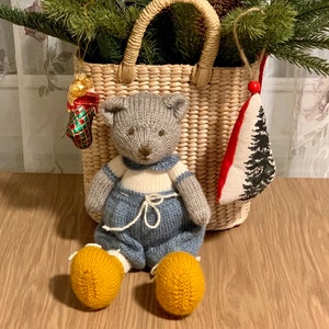 Könnte beinhalten: Ein grauer gestrickter Teddybär mit blauer Latzhose und gelben Schuhen sitzt vor einem Weidenkorb voller Weihnachtsbaumschmuck.
