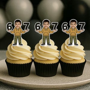 May include: Three cupcakes with yellow frosting and black cupcake liners are on a gray plate. Each cupcake is topped with a decorative figure of a boy holding the numbers 6 and 7. The background features black, white, and gray balloons.