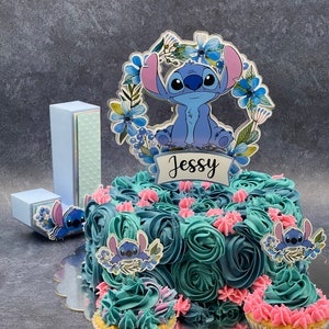 K&ouml;nnte beinhalten: Eine blau und t&uuml;rkis gefrostete Torte, verziert mit blauen und rosa Zuckerrosen und Stitch-Charakter-Toppern. Die Torte steht auf einem silbernen Tortenst&auml;nder und hat einen blauen und wei&szlig;en Pappbuchstaben 'J' sowie eine blaue und wei&szlig;e Pappbox mit einem Stitch-Charakter-Topper. Die Torte ist mit einem durchsichtigen Acryl-Topper mit einem Stitch-Charakter und dem Namen "Jessy" in Schwarz verziert.
