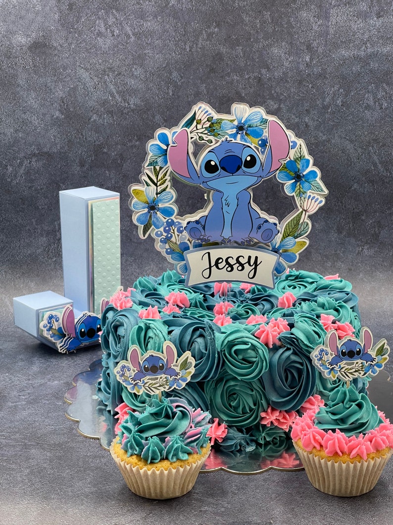 K&ouml;nnte beinhalten: Eine blau und t&uuml;rkis gefrostete Torte, verziert mit blauen und rosa Zuckerrosen und Stitch-Charakter-Toppern. Die Torte steht auf einem silbernen Tortenst&auml;nder und hat einen blauen und wei&szlig;en Pappbuchstaben 'J' sowie eine blaue und wei&szlig;e Pappbox mit einem Stitch-Charakter-Topper. Die Torte ist mit einem durchsichtigen Acryl-Topper mit einem Stitch-Charakter und dem Namen "Jessy" in Schwarz verziert.