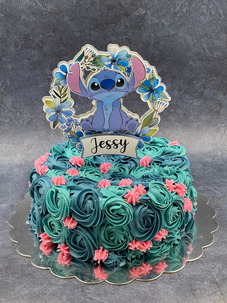 K&ouml;nnte beinhalten: Ein blauer und gr&uuml;ner Kuchen, der mit Buttercreme-Frosting in Form von Rosen verziert ist. Der Kuchen ist mit einer blauen und wei&szlig;en Figur aus dem Disney-Film Lilo & Stitch verziert. Der Kuchen steht auf einem silbernen Kuchenst&auml;nder und tr&auml;gt den Namen "Jessy".