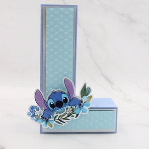 K&ouml;nnte beinhalten: Eine blaue und lila Comicfigur mit gro&szlig;en Augen, die hinter einem blauen und wei&szlig;en Blumenarrangement hervorlugt. Die Figur befindet sich auf einem blauen und wei&szlig;en Punktmuster. Die Figur ist der Buchstabe L.