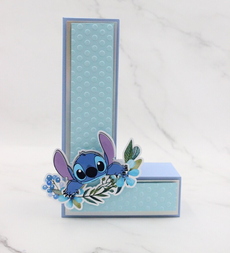 K&ouml;nnte beinhalten: Eine blaue und lila Comicfigur mit gro&szlig;en Augen, die hinter einem blauen und wei&szlig;en Blumenarrangement hervorlugt. Die Figur befindet sich auf einem blauen und wei&szlig;en Punktmuster. Die Figur ist der Buchstabe L.
