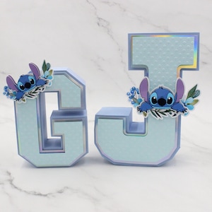 K&ouml;nnte beinhalten: Zwei hellblaue Buchstaben aus Pappe, G und J, verziert mit einer blauen Comicfigur und floralen Akzenten. Die Buchstaben haben eine strukturierte, gepunktete Oberfl&auml;che.