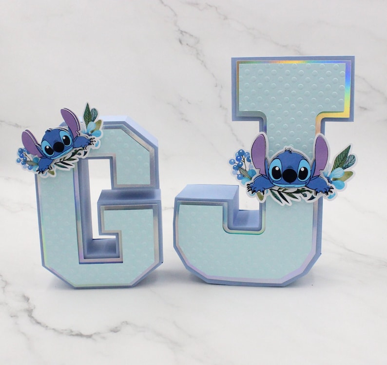K&ouml;nnte beinhalten: Zwei hellblaue Buchstaben aus Pappe, G und J, verziert mit einer blauen Comicfigur und floralen Akzenten. Die Buchstaben haben eine strukturierte, gepunktete Oberfl&auml;che.