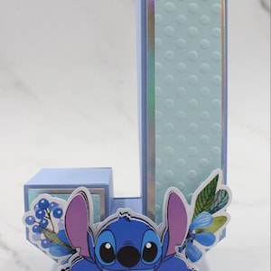 K&ouml;nnte beinhalten: Ein blauer und lila Stitch-Charakter, der hinter einem hellblauen Buchstaben J hervorlugt. Der Buchstabe ist mit blauen Blumen und gr&uuml;nen Bl&auml;ttern verziert. Der Hintergrund ist eine hellgraue Marmorfl&auml;che.