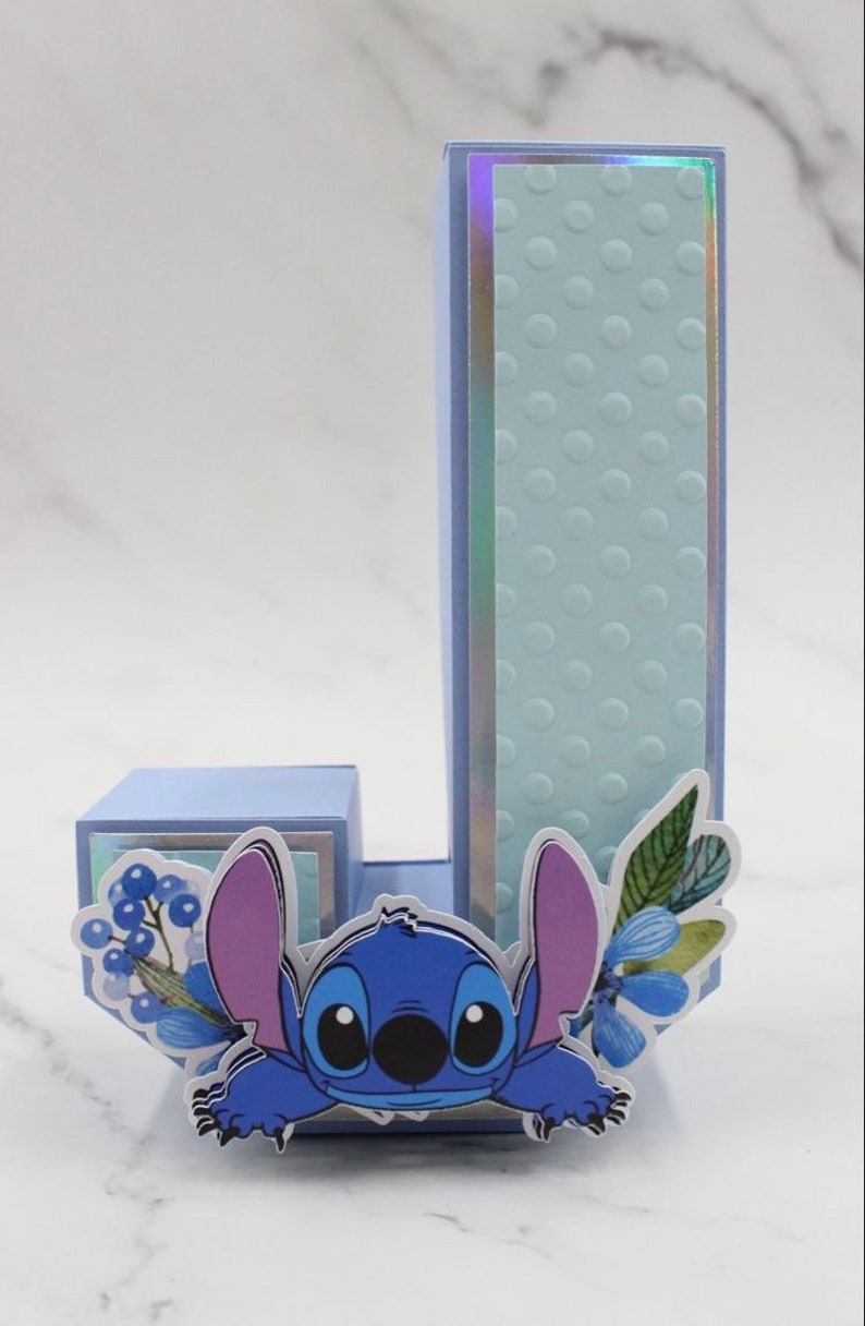 K&ouml;nnte beinhalten: Ein blauer und lila Stitch-Charakter, der hinter einem hellblauen Buchstaben J hervorlugt. Der Buchstabe ist mit blauen Blumen und gr&uuml;nen Bl&auml;ttern verziert. Der Hintergrund ist eine hellgraue Marmorfl&auml;che.