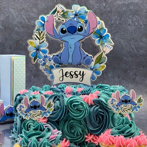 K&ouml;nnte beinhalten: Eine blaue und t&uuml;rkisfarbene Geburtstagstorte, verziert mit blauem und rosa Frosting und gekr&ouml;nt mit einem blauen Stitch-Charakter-Tortenaufsatz. Der Tortenaufsatz hat einen Blumengirlanden und den Namen "Jessy" darauf geschrieben. Es gibt zwei Cupcakes mit blauem und rosa Frosting und Stitch-Charakter-Tortenaufs&auml;tzen.