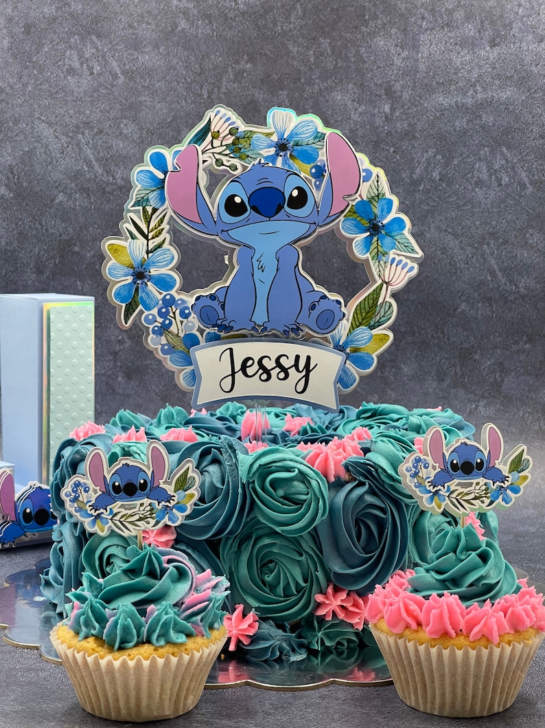 K&ouml;nnte beinhalten: Eine blaue und t&uuml;rkisfarbene Geburtstagstorte, verziert mit blauem und rosa Frosting und gekr&ouml;nt mit einem blauen Stitch-Charakter-Tortenaufsatz. Der Tortenaufsatz hat einen Blumengirlanden und den Namen "Jessy" darauf geschrieben. Es gibt zwei Cupcakes mit blauem und rosa Frosting und Stitch-Charakter-Tortenaufs&auml;tzen.