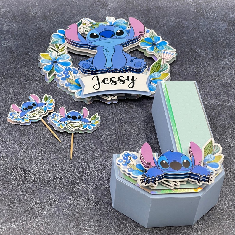 K&ouml;nnte beinhalten: Ein blauer und gr&uuml;ner 3D-Buchstaben-J-Kuchentopper mit einer blauen Stitch-Figur und floralen Akzenten. Der Topper tr&auml;gt die Aufschrift "Jessy" auf der Vorderseite.