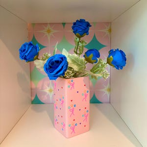 May include: A pink, square vase decorated with bows, stars, and hearts, displaying a bouquet of bright blue roses. The vase sits on a white shelf, against a backdrop of pink and teal geometric shapes.