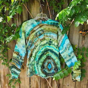 May include: A long-sleeved shirt with a tie-dye pattern in shades of blue, green, and brown. The shirt has a circular pattern with a darker blue center.
