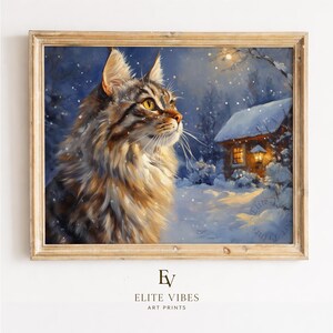 Op de afbeelding: Een ingelijste kunstdruk toont een langharige kat in een besneeuwde winterlandschap. De kat heeft bruine en bruinachtige vacht en gele ogen, kijkend naar een met sneeuw bedekt huisje met een verlicht raam. Het kunstwerk is gesigneerd "Elite Vibes Art Prints".