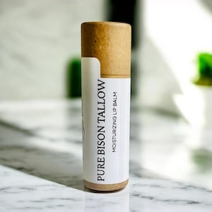 May include: A cylindrical lip balm tube with a light brown cardboard cap and base. The white label features the black text PURE BISON TALLOW and MOISTURIZING LIP BALM printed vertically. The container has a clean, minimalist design aesthetic.