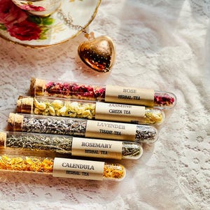 May include: Five clear glass test tubes with cork stoppers, each filled with dried botanicals and labeled with paper tags reading Rose Herbal Tea, Jasmine Green Tea, Lavender Tisane Tea, Rosemary Herbal Tea, and Calendula Herbal Tea.
