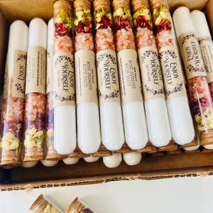 May include: A collection of glass test tubes filled with layers of white bath salts, pink Himalayan salt, and dried flower petals. Each tube is sealed with a cork stopper and features a paper label printed with the text Enjoy Yourself.