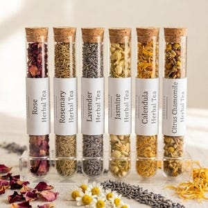 May include: Six glass tubes filled with various herbal teas, including Rose, Rosemary, Lavender, Jasmine, Calendula, and Citrus Chamomile. Each tube is labeled and sealed with a cork. The teas are displayed in a clear acrylic holder, with loose herbs and flowers scattered around.