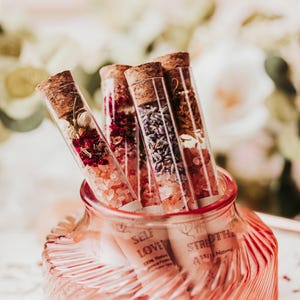 May include: Four clear glass test tubes with cork stoppers, filled with layers of pink salt and dried botanicals including lavender and rose petals. The tubes feature labels with text including SELF LOVE and STRENGTH, standing in a pink glass jar.