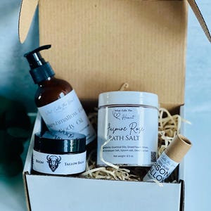 May include: A gift box containing body care products. Items include a brown bottle of body oil, a jar of jasmine rose bath salt, a small jar of tallow balm, and a lip balm. The products are packaged in a white box with straw-like filler.