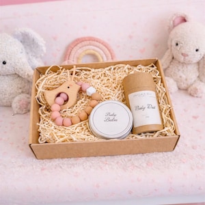 May include: A cardboard gift box filled with wood shavings containing a wooden bird-shaped teether with pink and tan silicone beads, a round metal tin labelled Baby Balm, and a cylindrical cardboard tube labelled Baby Dust in 2 oz size.