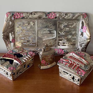 May include: A silver-toned decorative set featuring a rectangular tray with dragon and floral details, and two matching boxes with red and pink accents. A small, matching lighter is also present. The set is displayed on a wooden surface.