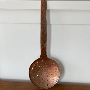 Puede incluir: Una cuchara ranurada de cobre vintage con un mango largo. El mango está decorado con un patrón repetitivo de medias lunas. La cuchara tiene un cuenco redondo con pequeños agujeros.