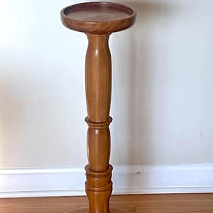 May include: A wooden plant stand with a round top and base, connected by a turned column. The stand is made of light-coloured wood and has a classic design. The stand is approximately 76 cm tall.
