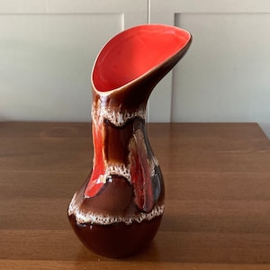 Puede incluir: Un jarrón de cerámica marrón y rojo con un diseño único y curvo. El jarrón tiene un acabado brillante y un interior rojo brillante.