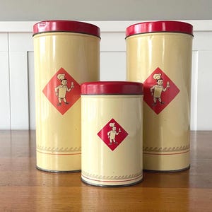 May include: Three vintage kitchen canisters in varying sizes. The canisters are cream-colored with red lids and bases. Each canister features a red diamond with a cartoon chef illustration. The canisters are arranged on a wooden surface.