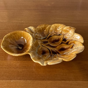 Op de afbeelding: Een bruine keramische serveerschaal met een bladontwerp en een kleine ronde kom eraan vastgemaakt. De schaal is geglazuurd in bruin en wit.