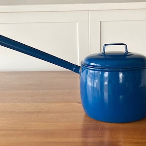 Peut inclure: Un arrosoir en émail bleu cobalt avec un long bec et un couvercle. La poignée est une simple boucle. Le pot est posé sur une surface en bois. Le design est simple et classique.