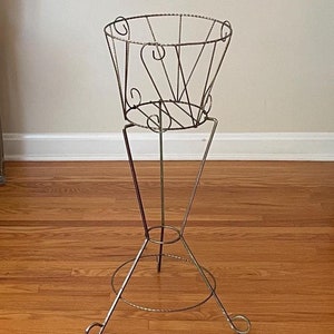 Puede incluir: Un soporte para plantas de metal dorado con una parte superior redonda y tres patas. El soporte está hecho de alambre y tiene un diseño decorativo.