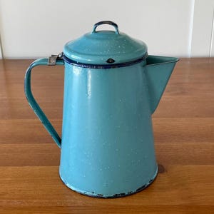 Peut inclure: Une cafetière en émail bleu vintage avec une finition mouchetée et un bord noir. La cafetière a un couvercle avec une petite poignée et un bec verseur sur le côté.