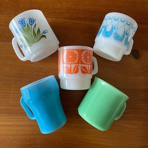 May include: Five vintage-style mugs in various colors and designs. The mugs include white with blue floral, white with blue teardrop, orange floral, blue, and green. Each mug has a handle and sits on a wooden surface.