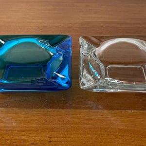 Puede incluir: Dos ceniceros cuadrados, uno de vidrio transparente y otro de vidrio azul vibrante. Los ceniceros tienen un diseño angular único, con una depresión central. Se muestran sobre una superficie de madera. Miden unos 10 cm.