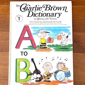 May include: The Charlie Brown Dictionary, Volume 1, by Charles M. Schulz. The cover features the letters A and B, with illustrations of Charlie Brown, Snoopy, and other Peanuts characters. The book is white with colorful illustrations.
