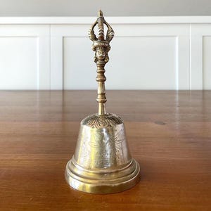 Könnte beinhalten: Eine Vintage-Messingglocke mit einem verzierten Griff und detaillierten Schnitzereien. Die Glocke hat einen ausgestellten Sockel und ein Blumenmuster, das in die Oberfläche geätzt ist. Der Griff hat eine dekorative Oberseite mit einem Gesicht und anderen komplizierten Designs. Die Glocke wird auf einer Holzoberfläche ausgestellt.